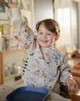 Smiling child in a Mushie Bluey Silicone Training Cup + Straw, seated at a table with a blue plate.