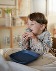 Smiling child in a mushie Long Sleeve Bib holds chopsticks at table with blue plate.