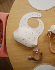 Mushie Silicone Baby Bib with pacifier and teether set displayed on a wooden table.