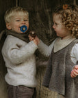 A young boy with a blue FRIGG pacifier and a girl with curly hair in cozy sweaters stand before a rustic wooden background. Smiling, the girl holds another FRIGG Natural Rubber 6-Pack pacifier—made in Denmark—near the boy’s face.