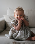 Baby in a plaid dress smiles on a couch, holding a mushie Teething Ring.
