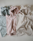 Three mushie Bear Poncho Towels in green, pink, and cream are laid out side by side. Each soft hooded towel features bear ears and fluffy fabric, displayed on a plain light background.