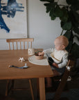 A baby with a mushie Figurine Teether sits at a wooden table in a cozy, modern dining area.