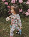 A young child in a floral outfit walks on grass holding a blue sippy cup, a beige bottle, and the mushie Linen Pacifier Clip 2-Pack, with pink rhododendron flowers and green leaves in the background.