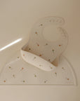 Cream-colored mushie Silicone Placemat and matching food-grade silicone baby bib with colorful rattle designs, shown on a light surface. The bib includes adjustable snap buttons and a practical food catcher pocket.