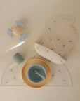 A flat lay of baby feeding items: a colorful silicone bib, yellow plate, blue cup and bowl with spoon, textured cup, mushie Silicone Placemat made from food-grade silicone, and a toy with clouds and sun on a white surface.