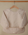 The mushie Long Sleeve Bib in light beige with small flower patterns hangs on a wooden hanger against a plain beige background. It features water resistance, elastic cuffs, a wide pocket, and a small white tag at the bottom.