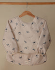 A light beige, water-resistant mushie Long Sleeve Bib with blue whale and fish prints hangs on a wooden hanger. It features elastic cuffs, a wide pocket, a white “mufle” tag, and is machine washable for easy cleanup.