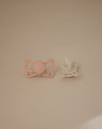 The FRIGG Butterfly Anatomical Silicone Pacifier 2-Pack (6-18 Months) by FRIGG features pastel pink and beige pacifiers with butterfly-shaped guards and anatomical nipples, arranged side by side on a light beige surface for healthy oral development.