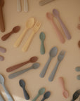 Mushie Dinnerware Fork and Spoon Set, dishwasher-safe, arranged on a beige surface flat lay.