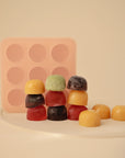 Red, yellow, green, and purple gummy candies are stacked in front of a pink Mushie Baby Food Freezer Tray with nine circular cavities, all set against a neutral background. The tray is made from BPA-free silicone.