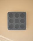 A Mushie Baby Food Freezer Tray, dark gray and square with nine round cavities, is centered on a beige surface. Soft light and shadows highlight its BPA-free silicone construction for safe baby food storage.