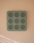 The Mushie Baby Food Freezer Tray is a square, green BPA-free silicone tray with nine round compartments, featuring a minimalist design perfect for storing baby food. It is shown on a light peach surface.