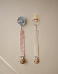 Two mushie Linen Pacifier Clips (2-Pack) with silicone clasps hang on a beige wall.