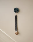 A mushie Linen Pacifier Clip from the 2-Pack set, featuring a dark gray linen strap, wooden end, and silicone clasp, hangs on a beige wall hook. The smooth background is softly lit with a diagonal shadow on the lower left side.