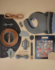 A flat lay in neutral tones showcases mushie's baby feeding essentials—bowls, spoons, bibs, placemat, teether, pacifier clip—alongside the Reusable Sticker Set with space-themed designs on a beige background.
