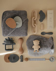 A neatly arranged flat lay of mushie baby bath and care items in neutral tones features towels, brushes, a No-Drop Dino Teether (Tradewinds), spoon, link chain, car toy, lotion box, and pacifier clip on a beige background.