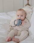 A baby in a light beige outfit sits on a white bed, holding and chewing on the mushie No-Drop Dino Teether (Tradewinds) in gray. White pillows and bedding are visible in the background.