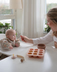 A smiling woman feeds a baby in a high chair at a white table. On the table is the Mushie Baby Food Freezer Tray and a small bowl. Sunlight streams through windows, with green plants on the windowsill.