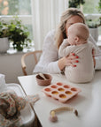 A woman with red nails holds a baby at a white table in soft natural light. On the table are a Mushie Baby Food Freezer Tray with puree, a divided bowl, and a toy. Green plants provide a fresh background.