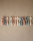 A row of mushie Silicone Toddler Starter Spoons, designed for independent feeding, displayed side by side on a plain tan surface in soft pastel shades.