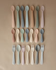 Mushie Silicone Toddler Starter Spoons in pastel shades are neatly arranged in rows on a neutral beige surface—perfect for encouraging independent feeding.