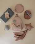 A flat lay of baby items, including a pink backpack, mushie’s Star Lovey Blanket in gray organic cotton, a pink star-shaped soft toy, a silicone pop-it toy, a divided pink lunchbox with utensils, a gray sippy cup, and a muslin cloth.