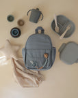 Flat lay of baby items on beige, including a light blue backpack, lunchbox with fork and spoon, sippy cup, stacking cups, mushie Star Lovey Blanket (organic cotton star-shaped lovey with cloth), and a pacifier clip.