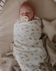 A baby sleeps peacefully in a cushioned bassinet, wearing a brown cap and using a pacifier, with the mushie Star Lovey Blanket and a soft floral-patterned swaddle beside them.