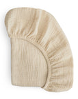 The mushie Extra Soft Bassinet Muslin Crib Sheet in beige features a textured design with elastic edges, partially folded on a white background—ideal for breathable and cozy baby bedding.