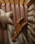 A wooden crib viewed from above in soft, warm light features a pink mattress, a star-shaped pillow, mushie’s cream Star Lovey Blanket made from organic cotton, and a giraffe plush toy resting on the bedding.