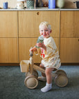 A toddler on a tricycle holds a mushie Soft Spout Sippy Cup in a cozy kitchen.