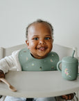 Smiling baby in a high chair wearing a green mushie Silicone Baby Bib, holding spoon and cup.