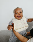 Smiling baby in mushie Silicone Baby Bib with adjustable neck strap, held by adult on gray couch.