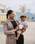 Smiling woman holding a baby and a mushie Snack Cup at an outdoor playground.