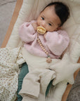 A baby in a pink knitted sweater and green pants lies in a cushioned bouncer, holding a beige pacifier clipped to their sweater, surrounded by soft blankets and the mushie Star Lovey Blanket.
