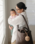 A woman in glasses and a white shirt holds a baby with the FRIGG Moon Natural Rubber Pacifier 2-Pack by FRIGG. She carries a large brown bag with baby essentials and a round keychain, standing by a window with blinds in soft light.