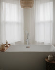 A modern white bathtub with a wooden tray holds mushie's Fragrance Free Baby Body Lotion, set in front of sheer white curtains and lit by natural light, with a woven pendant overhead and a small table beside the tub.