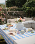 Outdoors, a picnic table with a blue and white striped cloth is set with mushie Dinnerware Fork and Spoon Set, teacups, plates, fruit, napkins, and flowers. Cushioned benches and lush greenery complete the scene.