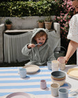 A child in a gray bear-ear robe sits outdoors, eating with a spoon. An adult in a white dress smiles beside them. The table features mushie Dinnerware Cups and pastel kids' dishes, with greenery visible in the background.