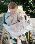 A toddler sits outdoors in a high chair, wearing a floral long-sleeve shirt, holding food. On the tray are a pink mushie Dinnerware Fork and Spoon Set plate with food and matching cup; wooden furniture and greenery are in the background.