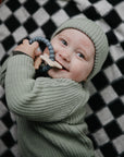 A smiling baby in a green knit outfit lies on a checkered blanket, holding the mushie Space Teething Ring—crafted from food-grade silicone with a star, ideal for sensory development.