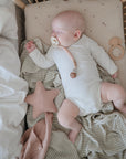 A baby in a white onesie sleeps in a crib on mushie’s Star Lovey Blanket, holding a pacifier. Nearby are a pink star-shaped pillow, teether, and cozy muslin cloth.