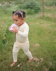 A young child wearing cream pajamas walks barefoot on the grass, sipping from a cup, with a mushie Linen Pacifier Clip 2-Pack attached to their outfit. Flowers and greenery create a lush garden background.
