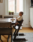 A young child sits in a high chair at the dining table, eating with a spoon. The Mushie Splat Mat protects the wooden floors, while potted plants on the windowsill soak up morning sunlight streaming through.