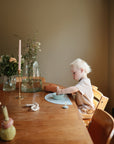 A young child with blond hair sits at a wooden table, playing with mushie Silicone Toddler Starter Spoons and a bowl. The table is adorned with candles, flowers in glass vases, and small items as sunlight filters through a nearby window.