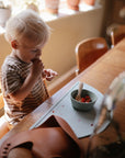 A young blond child in a striped shirt stands by a wooden table, eating from a small bowl with mushie Silicone Toddler Starter Spoons. Sunlight enters through a window, highlighting potted plants in the background.