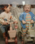 Two toddlers indoors on toy bikes, each holding a mushie Fairy Teething Ring.