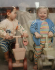 Two toddlers indoors on toy bikes, each holding a mushie Fairy Teething Ring.