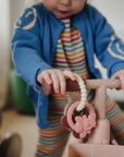 Toddler with mushie Fairy Teething Ring explores pink tricycle in striped outfit.